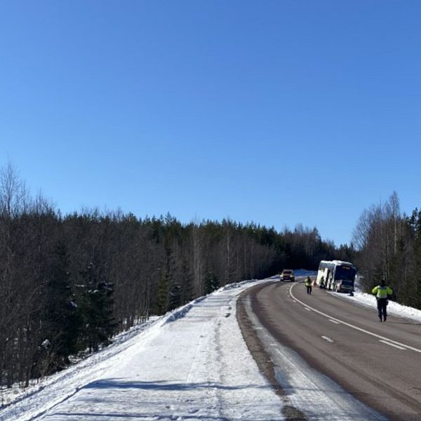 På bilden syns bussen i det högra hörnet, det är två personer som reflexvästar som går på vägen. Bilden är tagen utomhus och himlen är blå.
