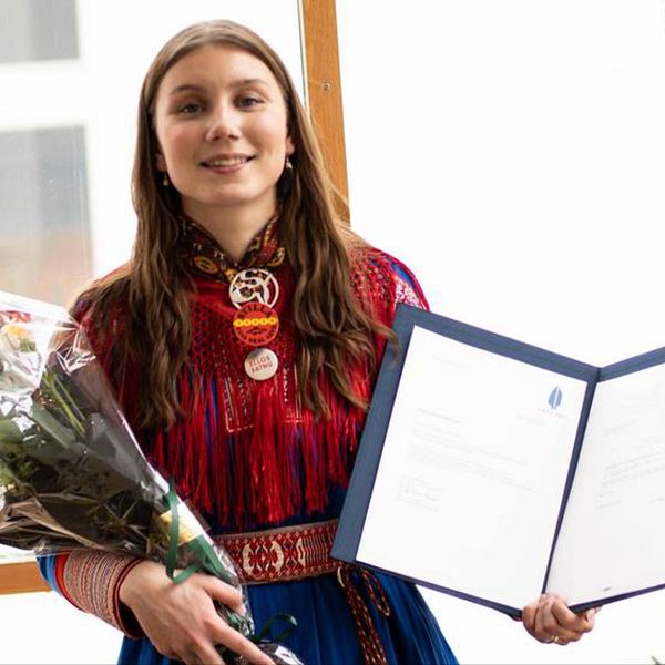 Ella Marie Hætta Isaksen som är en 25 årig tjej syns på bilden. Hon har långt brunt hår och bär på bilden en kolt från sitt samiska område. I ena handen håller hon en blombukett och i den andra en pärm med motiveringen till hennes vinst i den andra.