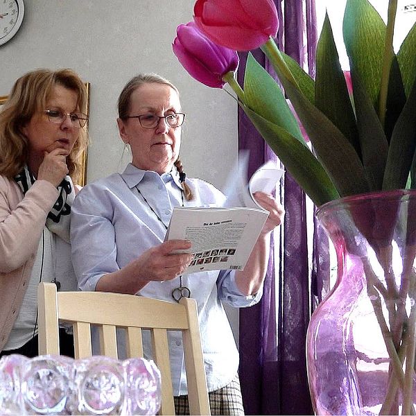 Två kvinnor står och läser i en broschyr. I förgrunden syns en vas med tulpaner.