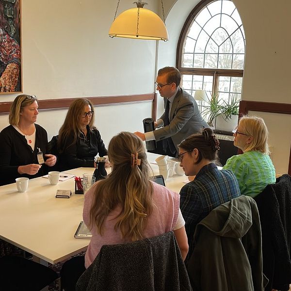 Sex kvinnor sitter samlade runt ett bord, socialborggarråd Alexander Ojanne (S) serverar kvinnorna kaffe.