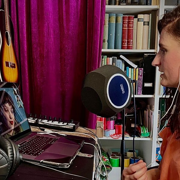 En tjej med rosa blus sitter frmaför en dator och har ett videosamtal. Hon har vita hörlurar i och en stor mikrofon framför sig.