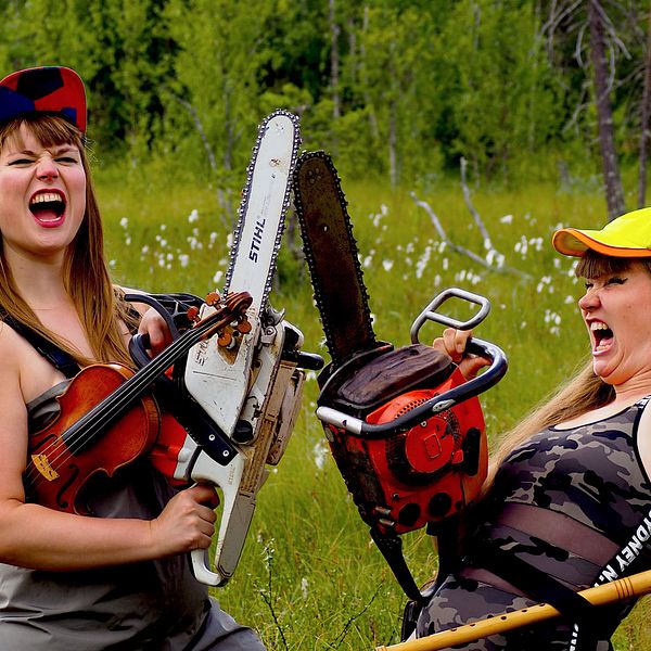Två tjejer i bandet Våtmark från Västerbotten står med varsin motorsåg och vadarbyxor i en skogsmiljö.