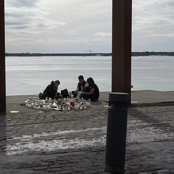Tre personer sitter vid minnesplatsen i Södra hamn. Tända ljus står på marken.