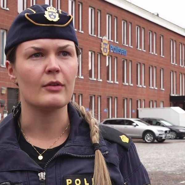 Kvinnlig polis framför Borlänge polishus. Hon har en polismössa med emblem på sig framför det tegelhuset.
