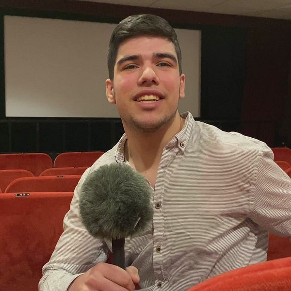en ung man sitter i en biosalong och pratar i en mikrofon, i bakgrunden syns filmduken