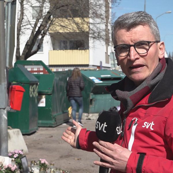 reporter framför återvinningsstation och blommor på marken