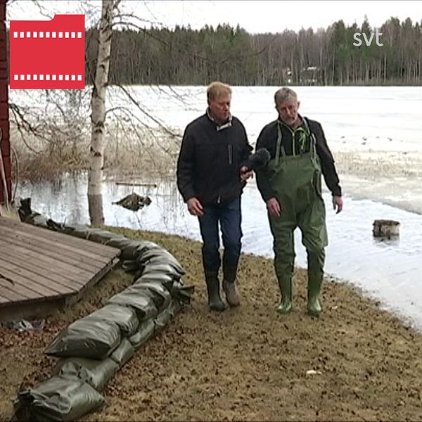 Två män går längs en rad med sandsäckar som ligger mellan en sjö och ett hus.