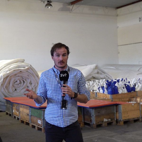 Reportern står framför ihopprullad folie och sandsäckar i ett stort lagerlokal.