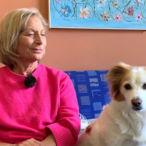 Birgitta Prytz sitter i en soffa framför en vägg med en hund till höger om henne som hon kollar ner på och ser glad ut.
