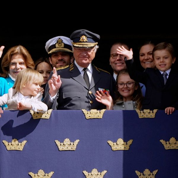 Kungen med familj och vänner vinkar från en balkong med kungliga symboler.