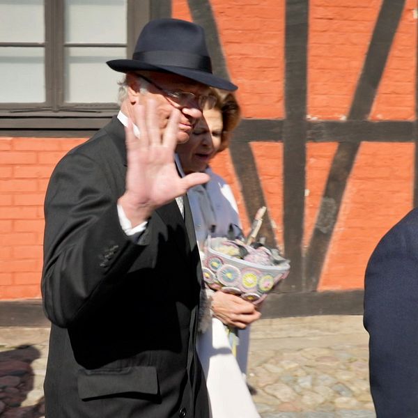 Kungen vinkar till kameran utomhus framför en röd tegelvägg.