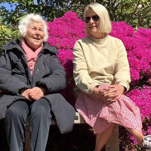 Två kvinnor, en mor och dotterm sitter på en parkbänk. Bakom dem är det fullt av rosa blommor på en buske.