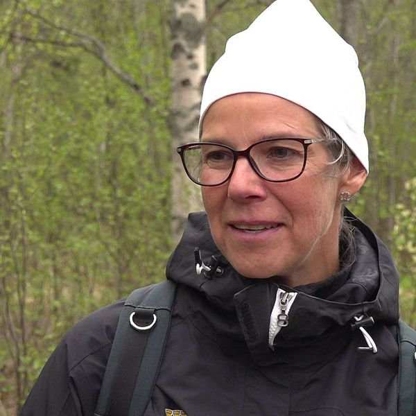 Kvinna med vit mössa, svart jacka och svartbågade glasögon står utomhus med grönskande skog bakom sig.