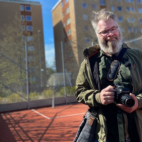 Man med skägg håller i kamera på en idrottsplan framför höghus i miljonprojektområde.