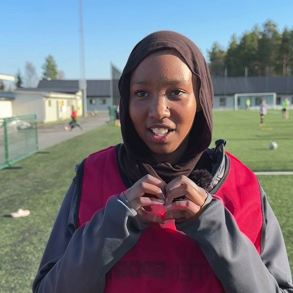 Najma Isse Hussein står framför en fotbollsplan och formar händerna till ett hjärta.