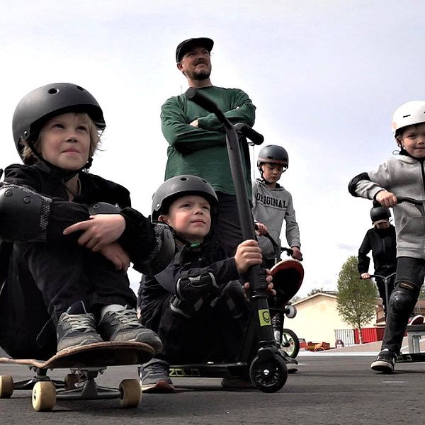 Barn sitter på sina skateboards och tittar förväntansfullt snett uppåt. De har hjälmar på huvudet.