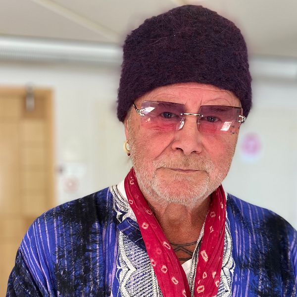 På bilden står Roger Pontare. Han har glasögon och en mörk mössa på sig. Han tittar in i kameran och har en blå/lila tröja och en röd halsduk.