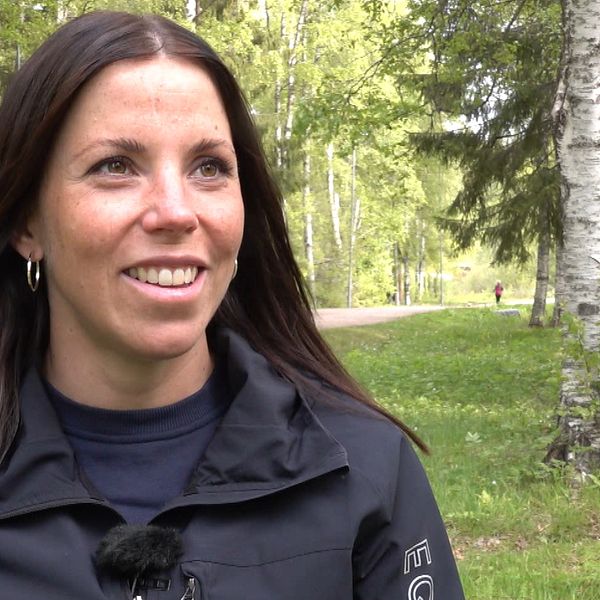 På bilden är Charlotte Kalla. Hon ler så att tänderna ses, hon har runda örhängen på sig och brunt hår. Hon tittar inte in i kameran och hon står vid gräs och träd.