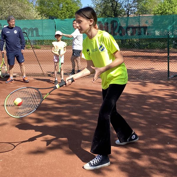 Ung tjej som spelar tennis samtidigt som några i bakgrunden tittar på.