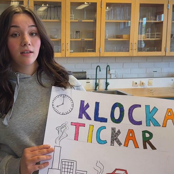 Amalia Lamnitchi står i ett klassrum på Gudmundråskolan i Kramfors. Hon har långt mörkt hår, grå huvtröja och håller i ett plakat med en klocka, industri och bil samt texten ”klockan tickar” i olika färger.