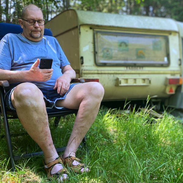 Bill Lundberg sitter i solstol utanför sin husbil och tittar i sin mobil.
