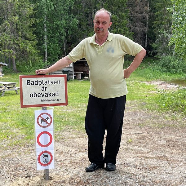 Man men gul skjorta håller handen på en skylt där det står ”Badplatsen är obevakad”