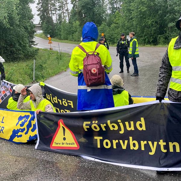 Ca sju personer i gul refelxvästar sitter på väg med banderoll märkt ”Återställ våtmarker. Torv är fossilt. Förbjud torvbrytning”. En person med Fjällräven-ryggsäck står med ryggen mot fotografen. I bakgruden står några poliser och talar med en person med reflexväst.