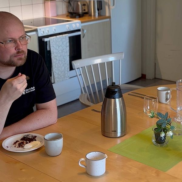 Anton Bylund har svarta kläder med Skellefteå kommuns logga på bröstet. Han sitter vid ett bord uppdukat med kaffekoppar med en bit kladdkaka i handen.