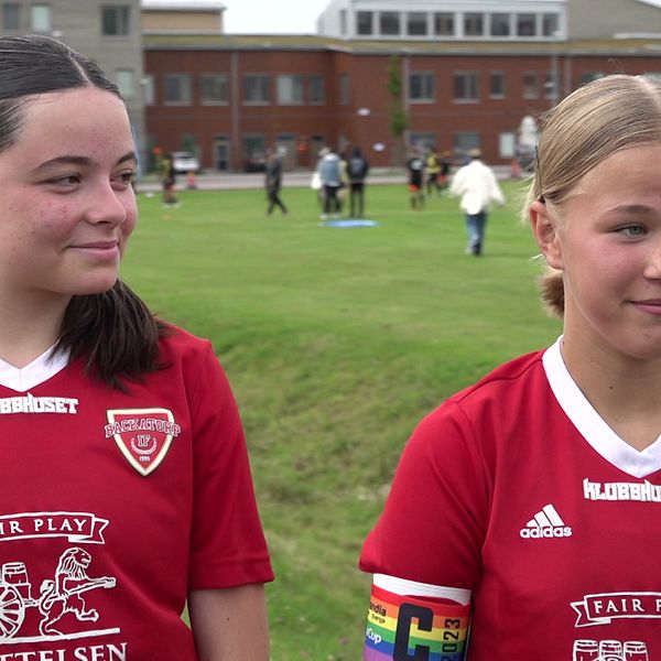 Två unga tjejer ses intervjuas, iklädda röda matchtröjor. I bakgrunden ser vi en fotbollsplan med besökare.