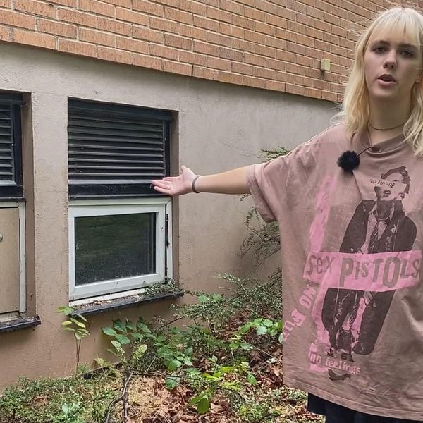 Blond kvinna tittar mot kameran och pekar mot två källarfönster varav ett är förspikat. Lila t-shirt med Sex Pistols på. Dea Nilsson, Falkvikskyrkan, Sölvesborg, Kyrkpunk.