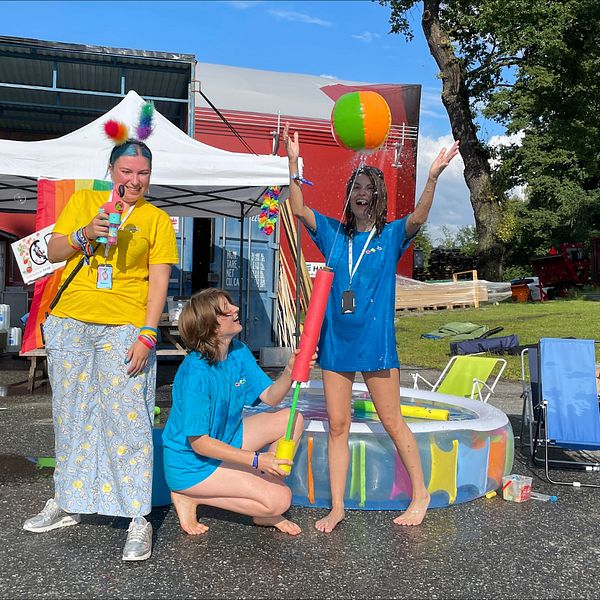 Lizzie, Nora och Stella jobbar som volontärer på Stockholm Pride och har hjälpt till att bygga upp Pride Park på Östermalms IP. På bilden sprutar volontärerna med vattenpistoler och kastar boll vid en barnpool i regnbågsfärger som är placerad vid byggboden där allt materiel och verktyg förvaras. Volontärerna skrattar.