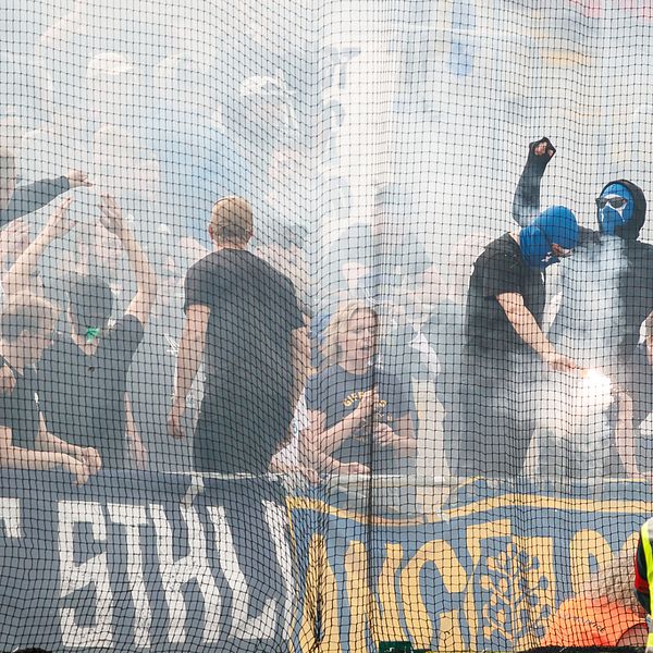 Flera maskerade supportrar står på läktaren under fotbollsderbyt mellan Östersunds FK och Gif Sundsvall i juli 2023. Runt dem virvlar rök från pyroteknik.