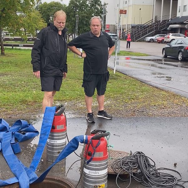 Två män står vid en brunn på en väg vid Timrå Arena och jobbar med översvämningen.