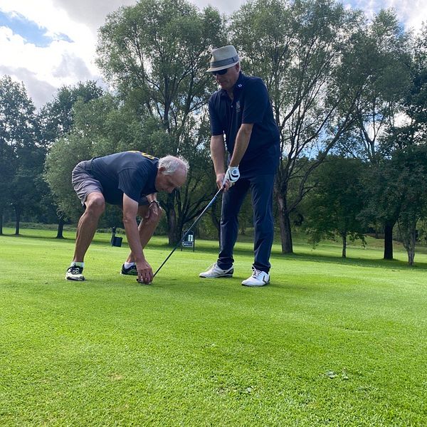 Två män spelar golf, en caddie visar en synskadad man hur han ska placera klubben för att få en bra träff