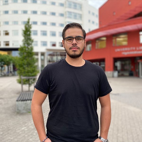 Bild på en ung kille i svart t-shirt och glasögon framför högskolan.