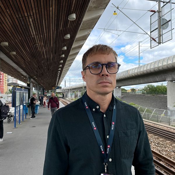 En man i glasögon och blå tröja som ser allvarlig ut på en pendeltågsstation