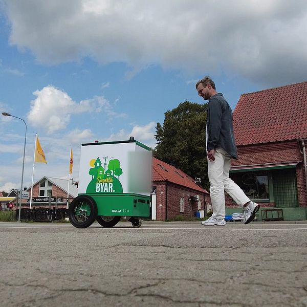 En självkörande robot formad som en låda på cykelbanan i Veberöd. På roboten står Smarta byar. Bakom roboten går produktdesigner Anton Ebel.