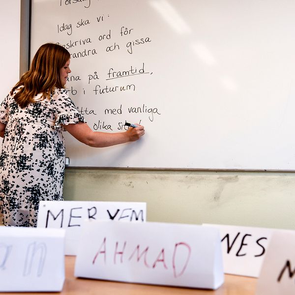 En kvinna i klänning skriver på en whiteboard. Framför henne syns namnlappar.