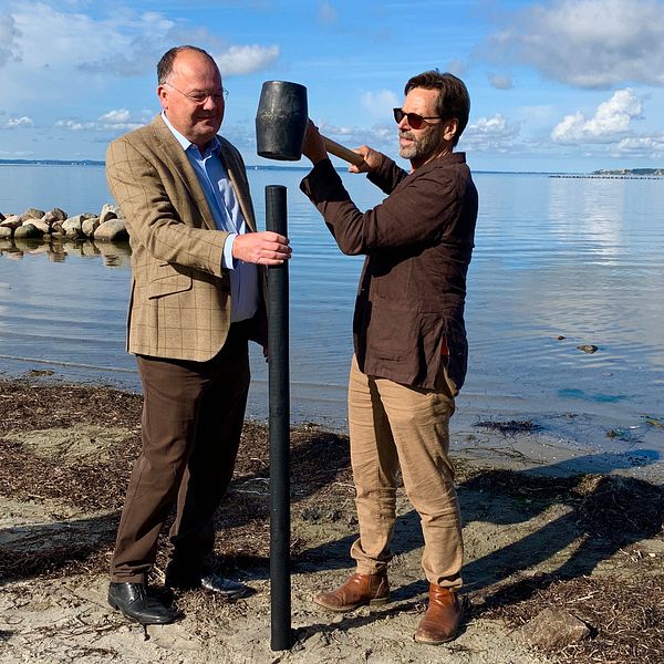 Kommunstyrelsens ordförande i Landskrona, Torkild Strandberg (L) och kallbadarföreningen Doppingarnas ordförande, Kjell Svegrup står vid havet i Landskrona. Där slår de gemensamt ned en påle i stranden som symboliserar det först ”spadtaget”.