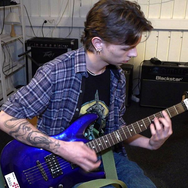 Ung man med mörkt hår och tatueringar spelar på en blå elgitarr.