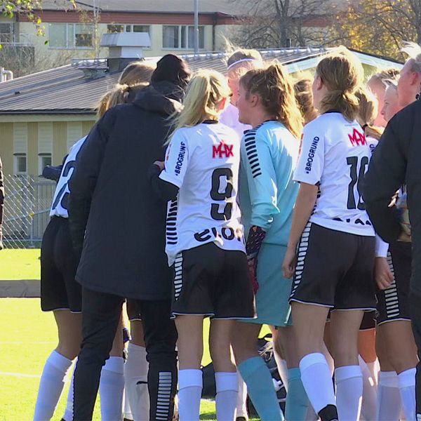 Unga tjejer som spelar fotboll i vita tröjor står i en ring. På höger och vänster sida syns tränare i svarta jackor.
