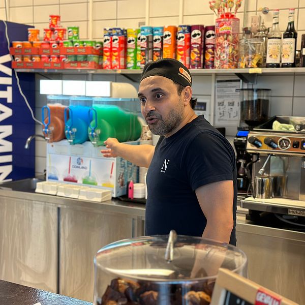 Kioskägaren Aghil Erfani Kia står vid kassan och pekar in mot kiosken, i bakgrunden syns olika sorters chips och en slushimaskin.
