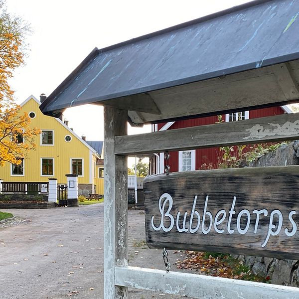 Skylt på Bubbetorps gård med vit text, i bakgrunden ett rött och ett gult hus samt ett höstfärgat träd.