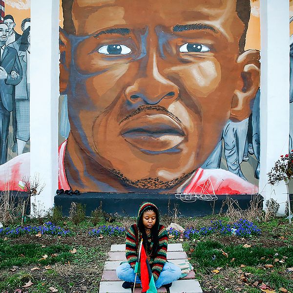 En kvinna sitter framför en väggmålning som föreställer Freddie Gray och som satts upp intill platsen där han greps.