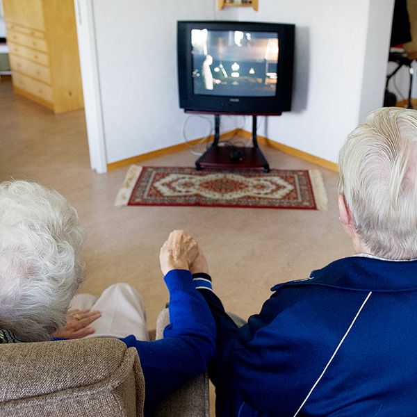 Ett äldre par håller varandra i handen och tittar på tv i sin lägenhet.