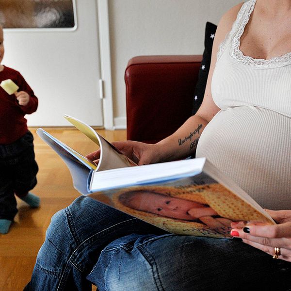 Gravid kvinna läser bok medan litet barn tittar på.