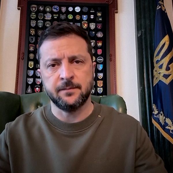 Ukrainas president Volodymyr Zelenskyj sitter vid sitt skrivbord med ukrainska flaggor bakom sig.