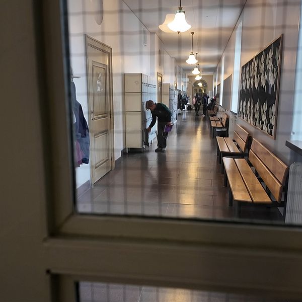 Elever i en skolhall syns genom ett glasfönster, några står vid skåpen.
