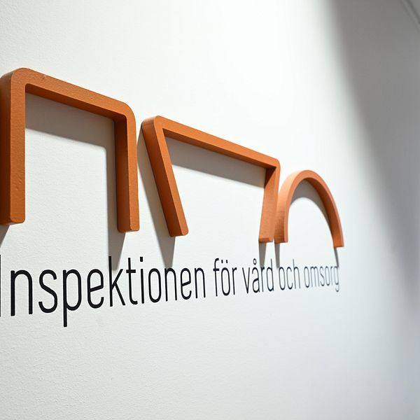 Logotypen för Inspektionen för vård och omsorg, med stiliserade bokstäver i orange och svart, placerad på en vägg i ett kontorsutrymme.