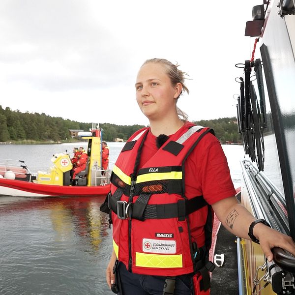 Sjöräddaren Ella Sjöberg i flytväst, står vid en båt med en räddningsbåt i bakgrunden.